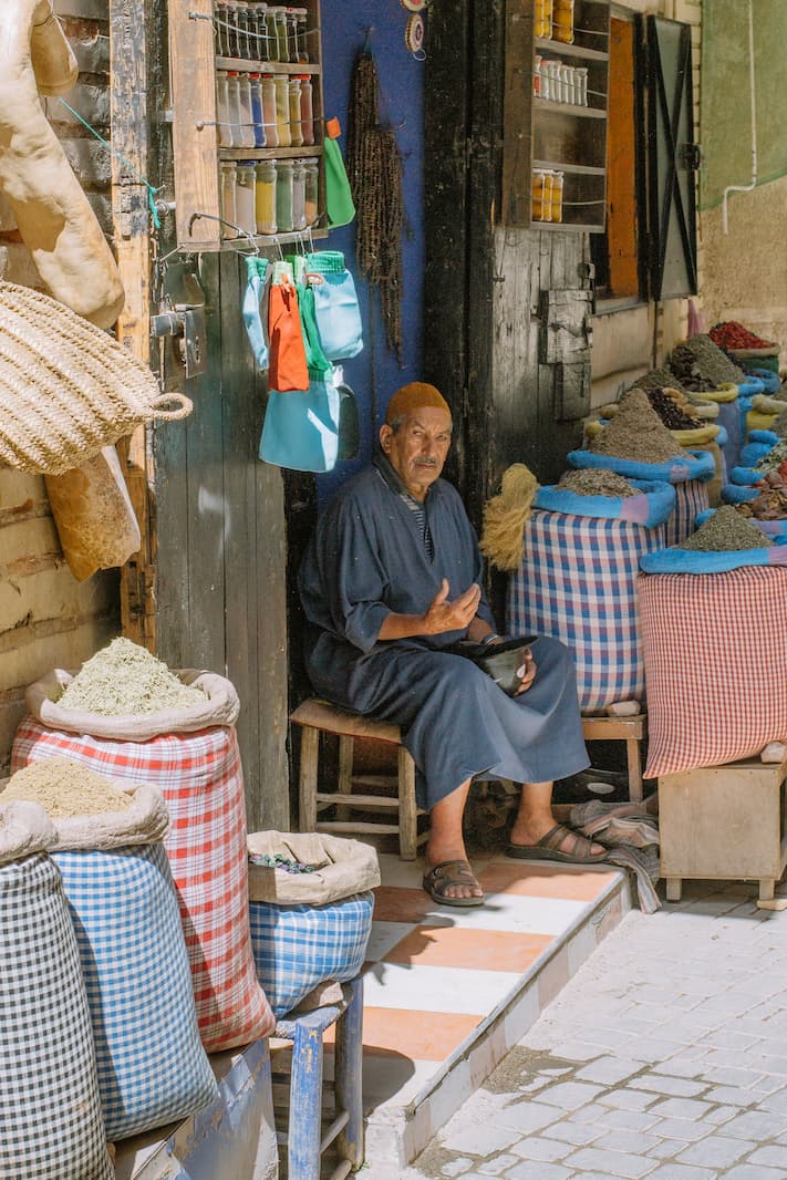 A man sits beside large bags of spices in a market