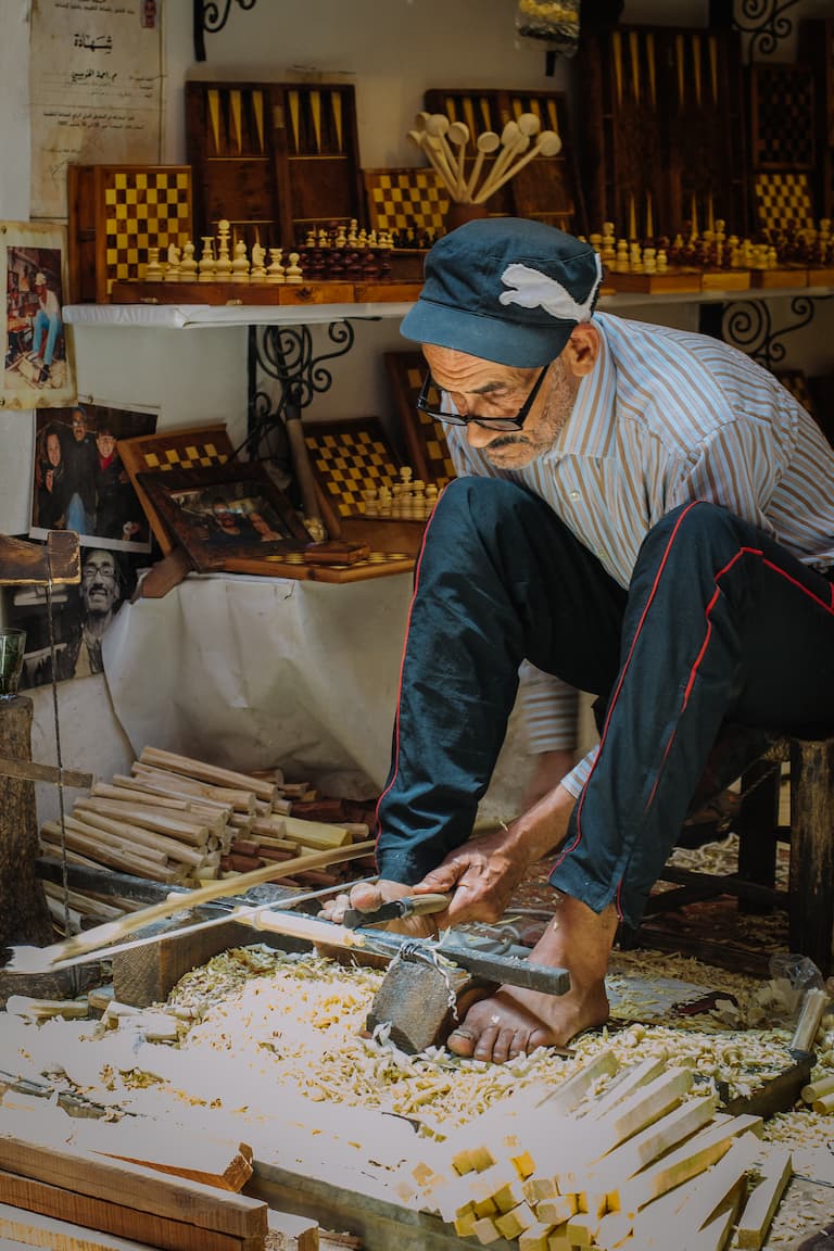 A man in a market uses using a homemade lathe to create chess pieces. He uses one hand to turn the lathe, the other to hold the carving tool and his foot to guide it along the wood's surface