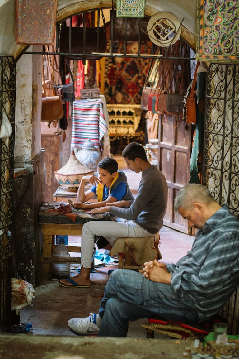 A father sleeping at the entrance to his shop. Inside his children sit at a table 