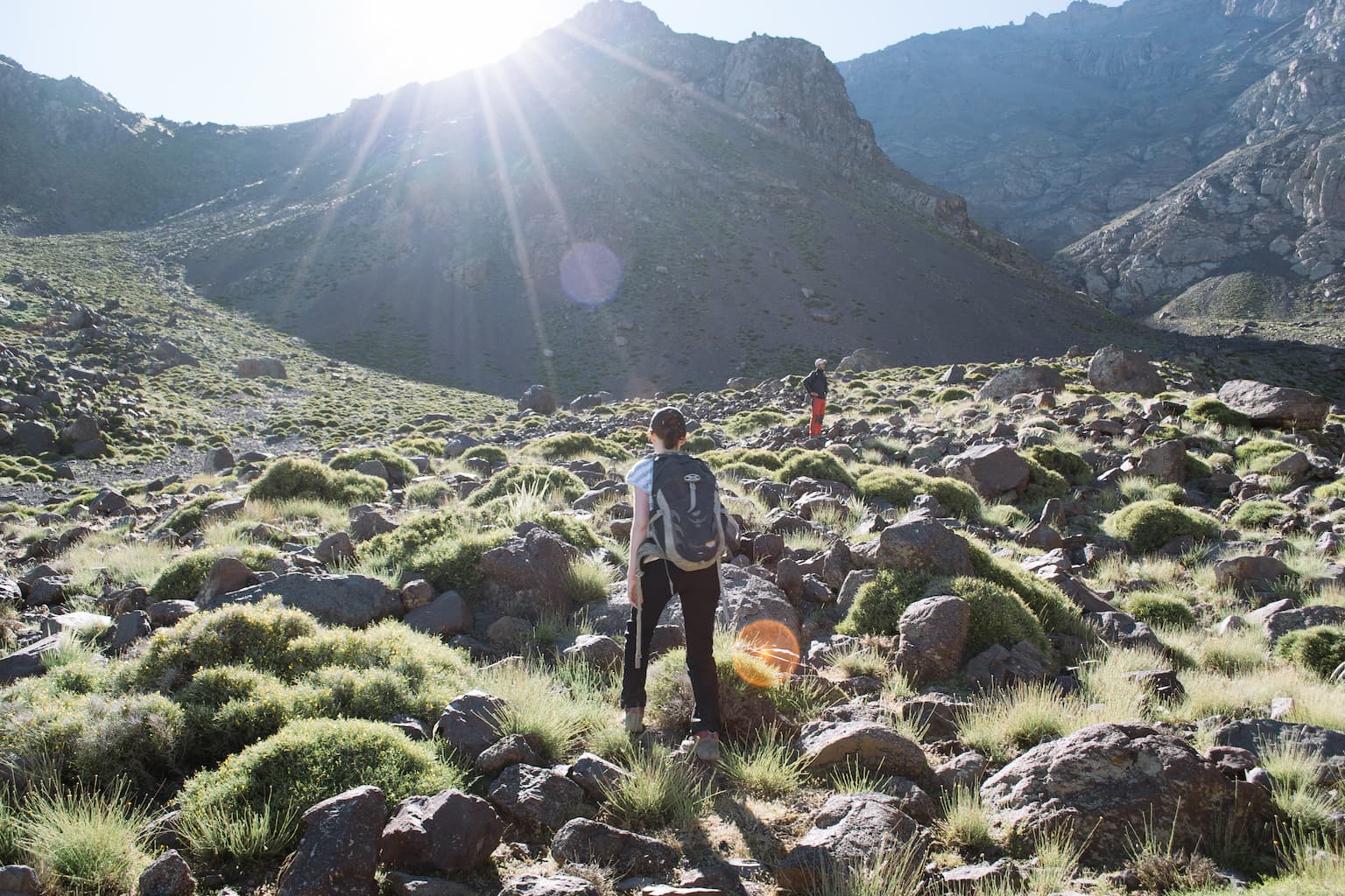 Sarah walking through a boulder field with the sun breaking over a hill in the distance