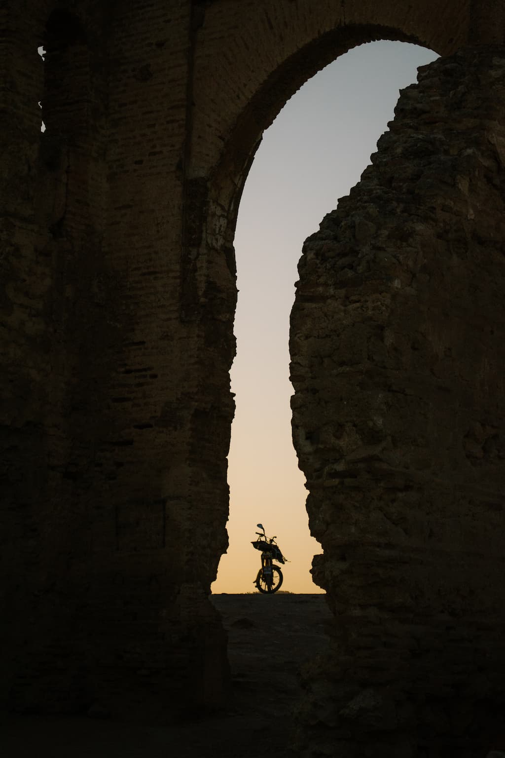 A gap in an old building shows a motorbike silhouetted against an orange sky